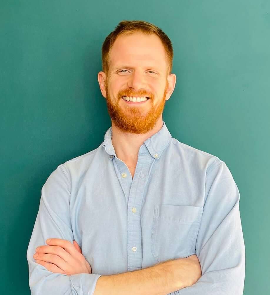 Dr. Alex Johnson is a fit looking man with red hair and smiling. He is standing against a teal colored wall